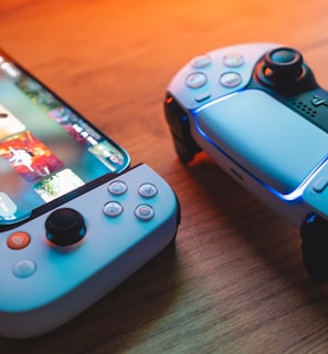 A gaming scenario featuring a smartphone with a game interface next to two controllers on a wooden surface. The smartphone displays vibrant images and icons, while the controllers feature a sleek, modern design with buttons and joysticks prominently visible. Warm lighting enhances the scene.